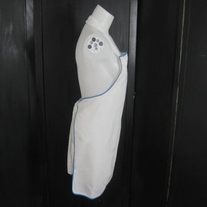 May include: White apron with a blue trim. The apron is hanging on a mannequin.