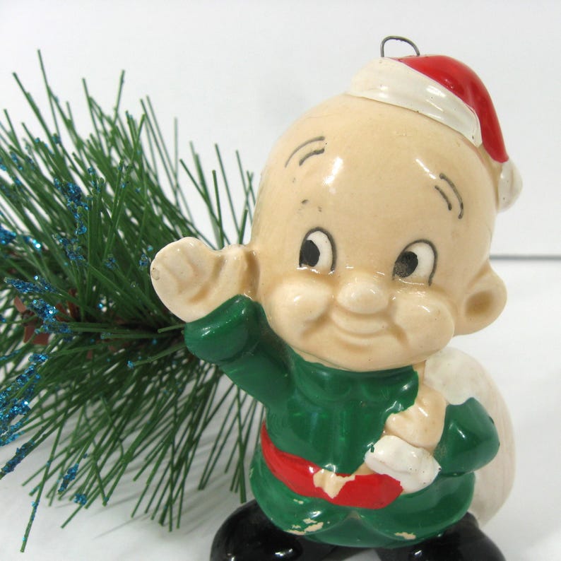 May include: A vintage ceramic Christmas bauble featuring a cartoon character dressed in a green suit, red hat, and carrying a sack. The character has a bald head, large eyes, and a wide smile.