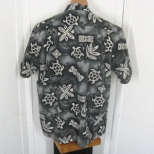 May include: A short-sleeved button-down shirt in shades of gray, black, and white. The fabric features a repeating pattern of sea turtles, stylized floral shapes, and geometric tribal-inspired motifs across the entire garment surface.