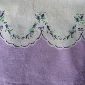 May include: A vintage textile with a scalloped edge, featuring a lavender lower section and a white upper section. The white section is adorned with a repeating pattern of purple floral bouquets and green leaves, accented with purple bows. The fabric has a soft, textured appearance.