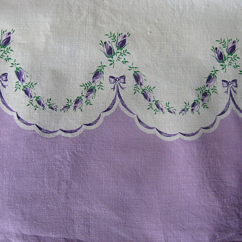 May include: A vintage textile with a scalloped edge, featuring a lavender lower section and a white upper section. The white section is adorned with a repeating pattern of purple floral bouquets and green leaves, accented with purple bows. The fabric has a soft, textured appearance.