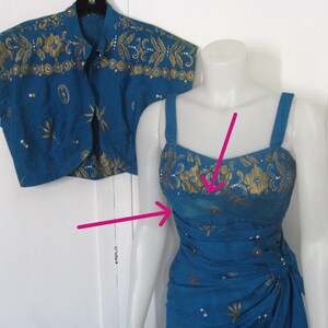 May include: A teal two-piece outfit comprising a sleeveless dress and a matching bolero jacket. The dress has a fitted bodice and a draped skirt. Both items are decorated with gold floral and geometric patterns.