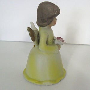 May include: A ceramic angel figurine in a yellow dress, holding a pink flower. The angel has brown hair and small, gold-tipped wings. The figurine is approximately 4 inches tall.