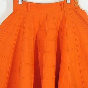 May include: A bright orange skirt with a box pleat design. The skirt is made of a textured fabric and has a high waist.
