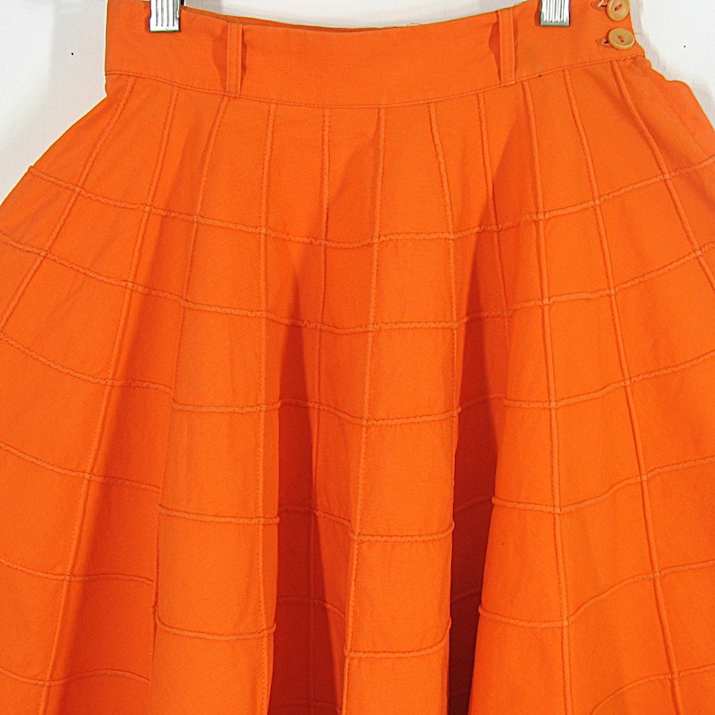 May include: A bright orange skirt with a box pleat design. The skirt is made of a textured fabric and has a high waist.