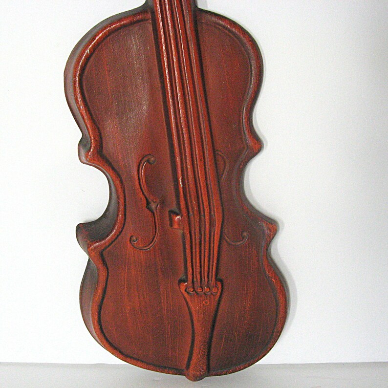 K&ouml;nnte beinhalten: Ein dunkelbrauner Wandbehang aus Holz in Form einer Geige. Das St&uuml;ck zeigt geschnitzte Details wie den Korpus, den Hals, die Saiten und die F-L&ouml;cher. Die Oberfl&auml;che weist eine sichtbare Holzmaserung mit matter Optik und abgerundeten Kanten auf.
