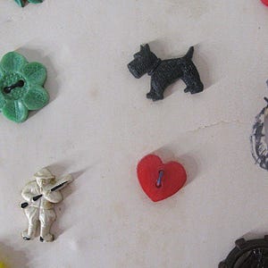 May include: A collection of assorted buttons including a green flower-shaped button, a black dog-shaped button, a white figure of a person holding a rifle, a red heart-shaped button, and a dark circular gear-shaped button with a ship design.