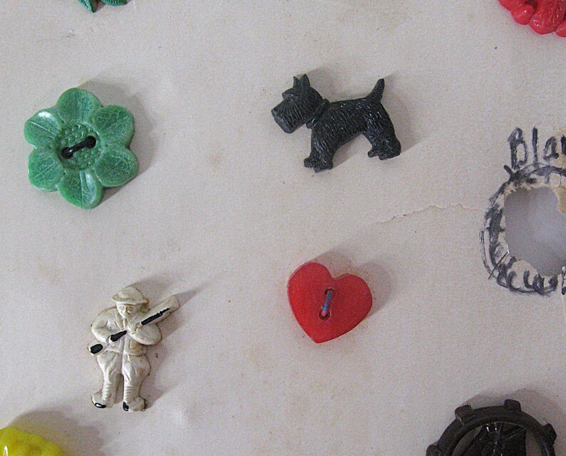 May include: A collection of assorted buttons including a green flower-shaped button, a black dog-shaped button, a white figure of a person holding a rifle, a red heart-shaped button, and a dark circular gear-shaped button with a ship design.