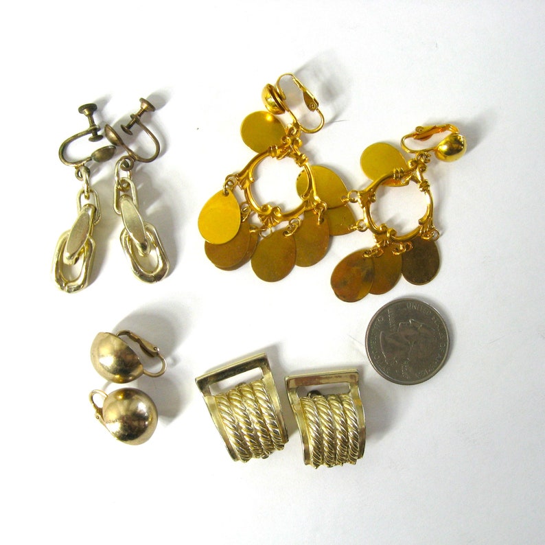 K&ouml;nnte beinhalten: Sortiment an goldfarbenen Ohrclips. Die Ohrringe haben verschiedene Stile, darunter ein Paar mit baumelnden m&uuml;nzf&ouml;rmigen Anh&auml;ngern, ein Paar mit einem seilartigen Design und ein Paar mit runden Kugeln.