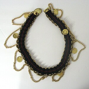 May include: A black fabric necklace with gold-colored coins and chains. The necklace has a red stitched detail down the center and a gold clasp. The coins are evenly spaced around the necklace, with chains hanging from them.