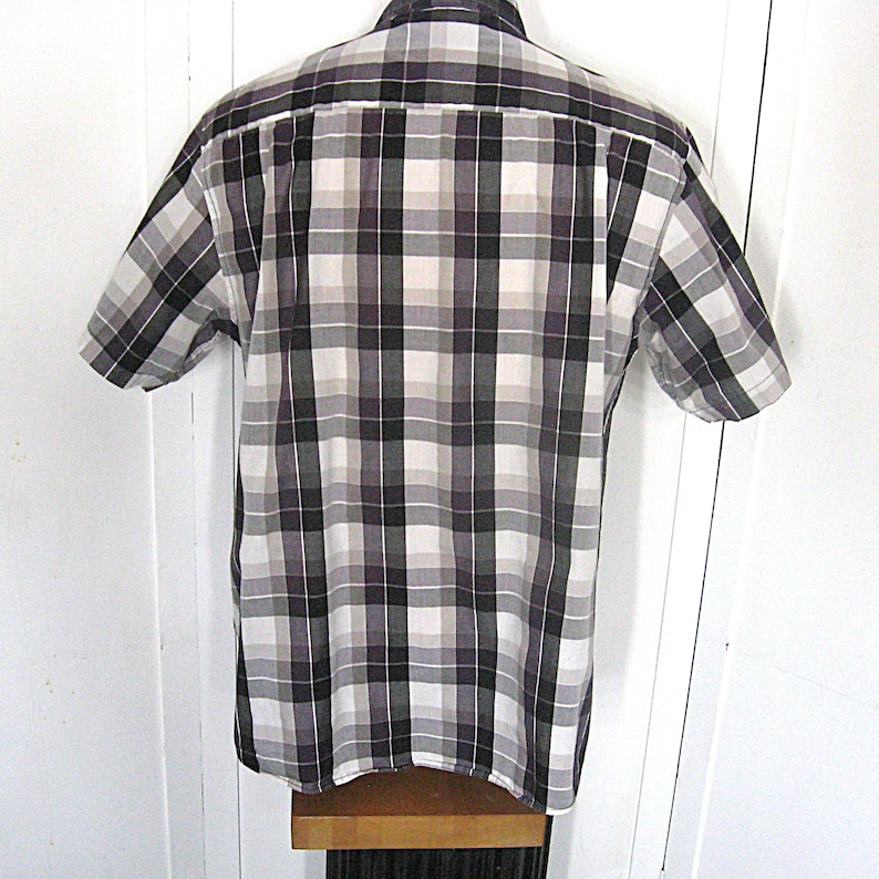 May include: Short-sleeved plaid shirt in a neutral color palette of black, white, and shades of gray. The shirt has a classic button-down design with a collar and short sleeves. The plaid pattern is evenly spaced.