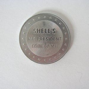 May include: A silver coin featuring the text "SHELL'S MR. PRESIDENT COIN GAME". The coin has concentric circles in the centre and is edged with small star shapes. A vintage collectible.