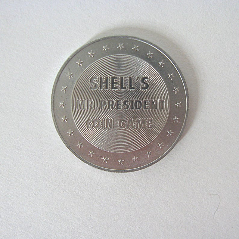 May include: A silver coin featuring the text "SHELL'S MR. PRESIDENT COIN GAME". The coin has concentric circles in the centre and is edged with small star shapes. A vintage collectible.