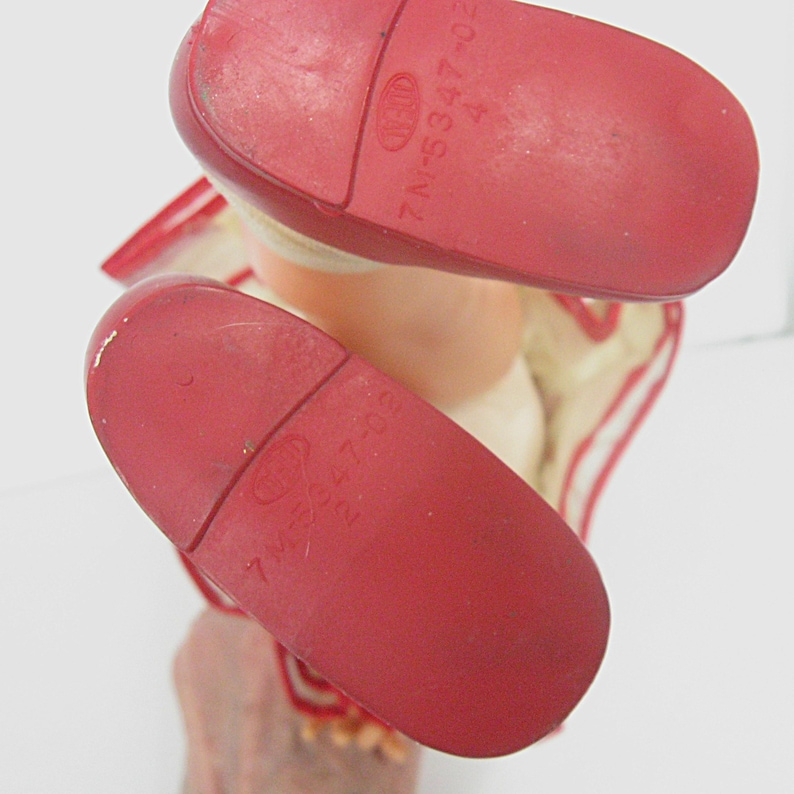 May include: The soles of a pair of red doll shoes, each featuring a molded logo and the embossed text 7M-5347-02. The shoes are attached to light-colored doll legs, with red trim visible around the ankle area of the footwear.