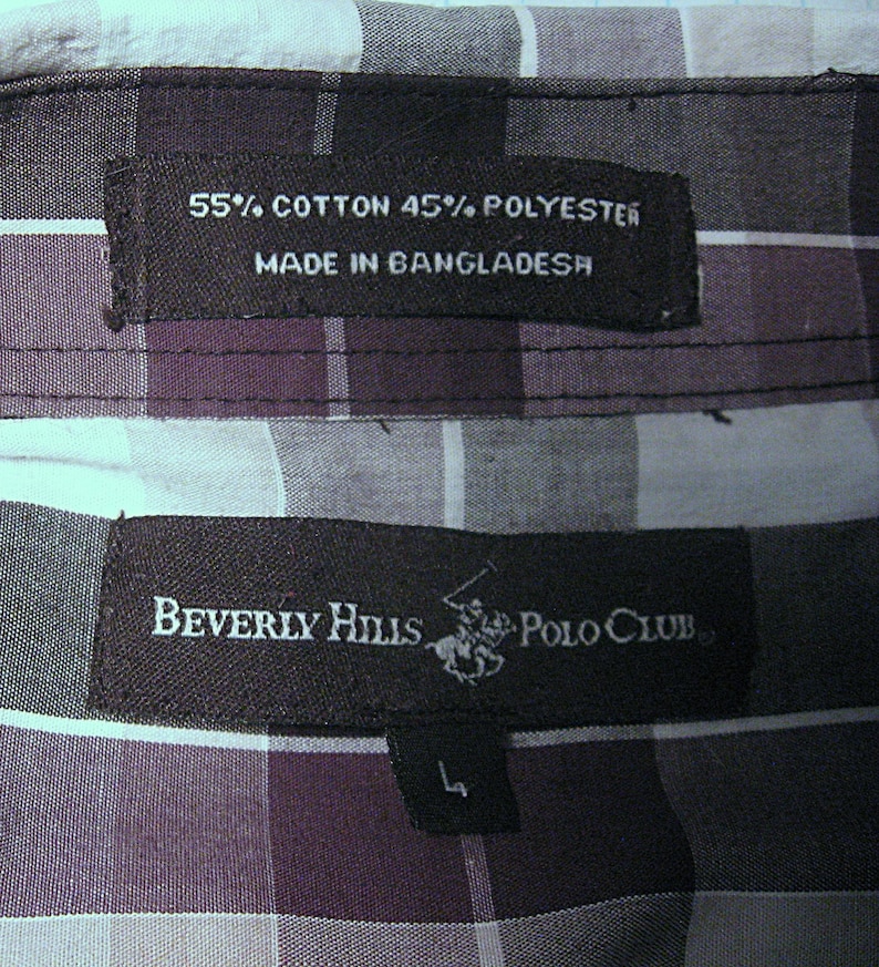 May include: Close-up of a plaid shirt label. The label reads "55% Cotton 45% Polyester Made in Bangladesh." Another label reads "Beverly Hills Polo Club" with a logo and size "L". The shirt has a purple, black, and white plaid pattern.