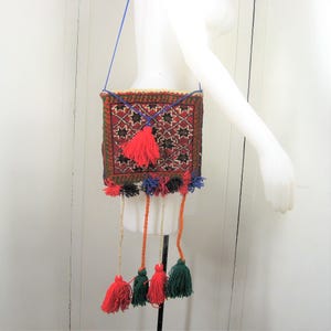 May include: A square, woven shoulder bag with a geometric pattern in red, black, and green. The bag has a blue strap and is adorned with red, green, and orange tassels. The bag is displayed on a mannequin.