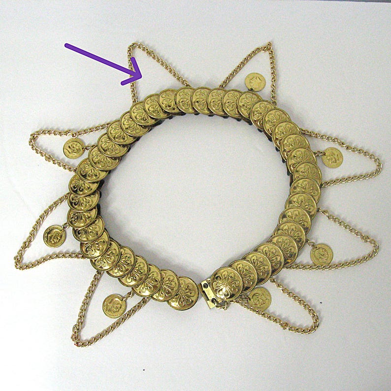 May include: A gold-colored necklace with a circular design, featuring numerous coin-like embellishments. The necklace has a star-shaped design with dangling chains and small coin pendants. The clasp is visible at the front.