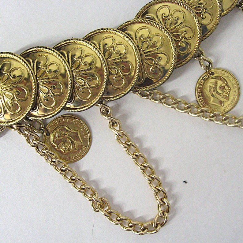 May include: A gold-toned belt featuring large, round, decorative medallions with an ornate floral design. The belt includes dangling coin charms and chain links, creating a vintage aesthetic. The belt is a statement piece.