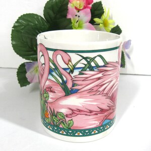 May include: A white ceramic mug featuring a pink flamingo design. The mug depicts two pink flamingos with green foliage and a blue water background. The mug has a green and white border with a geometric pattern.