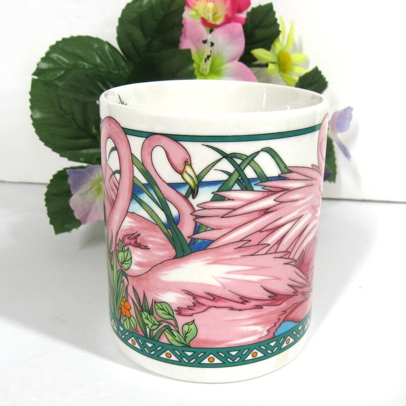 May include: A white ceramic mug featuring a pink flamingo design. The mug depicts two pink flamingos with green foliage and a blue water background. The mug has a green and white border with a geometric pattern.
