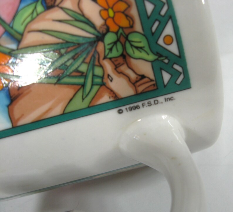 May include: White ceramic mug with a green, orange and brown design. The mug has a handle and the text "&copy; 1996 F.S.D., Inc." on the bottom.
