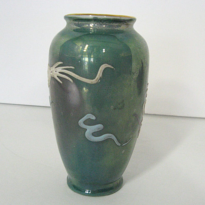 Pode incluir: Um vaso de cer&acirc;mica verde com acabamento brilhante. O vaso apresenta desenhos brancos estilizados de drag&otilde;es ou serpentes. O vaso tem um corpo arredondado e uma borda ligeiramente alargada. O design geral &eacute; simples e elegante.