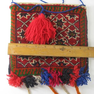 May include: A rectangular, woven bag with a red background and geometric patterns in black, white, and green. The bag has a red tassel and colorful tassels along the bottom. The bag is approximately 9 inches wide.