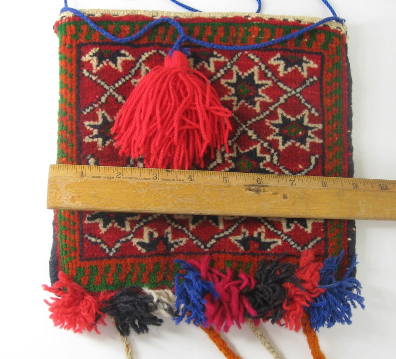 May include: A rectangular, woven bag with a red background and geometric patterns in black, white, and green. The bag has a red tassel and colorful tassels along the bottom. The bag is approximately 9 inches wide.