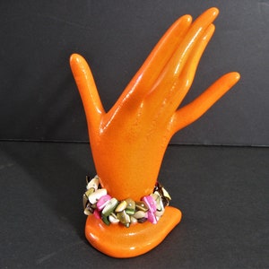 May include: A colorful bracelet made of various shades of brown, pink, and green shell pieces. The bracelet is displayed on an orange hand-shaped stand.