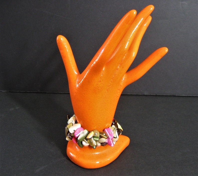 May include: A colorful bracelet made of various shades of brown, pink, and green shell pieces. The bracelet is displayed on an orange hand-shaped stand.
