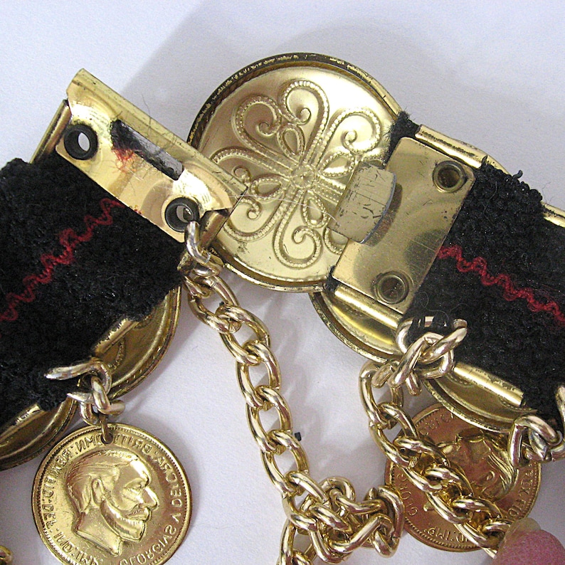 May include: Close-up of a vintage belt featuring gold-toned metal hardware and a black fabric strap with red stitching. The belt includes decorative gold coins and a chain. The buckle has an ornate floral design.
