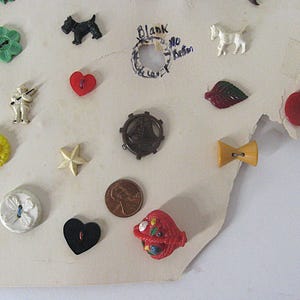 May include: A collection of assorted vintage buttons attached to a cream-colored card, including shapes like a green flower, black dog, white horse, red heart, yellow star, brown ship wheel, red apple, and a red basket, alongside a copper penny.