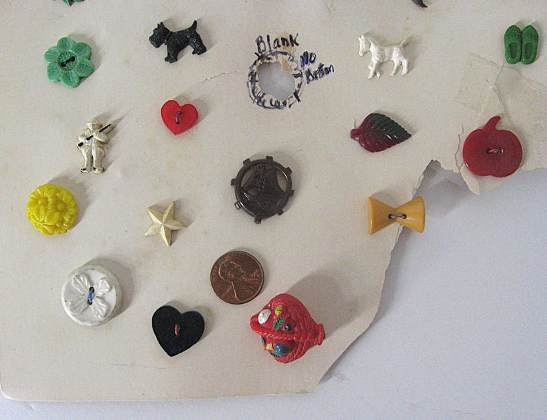 May include: A collection of assorted vintage buttons attached to a cream-colored card, including shapes like a green flower, black dog, white horse, red heart, yellow star, brown ship wheel, red apple, and a red basket, alongside a copper penny.