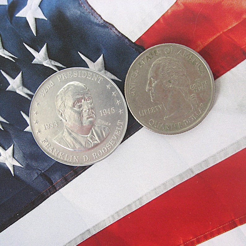 May include: Two coins on an American flag: a silver commemorative coin with Franklin D. Roosevelt's portrait and a quarter. The flag has red and white stripes and a blue section with white stars. The coin features the text "Quarter Dollar".