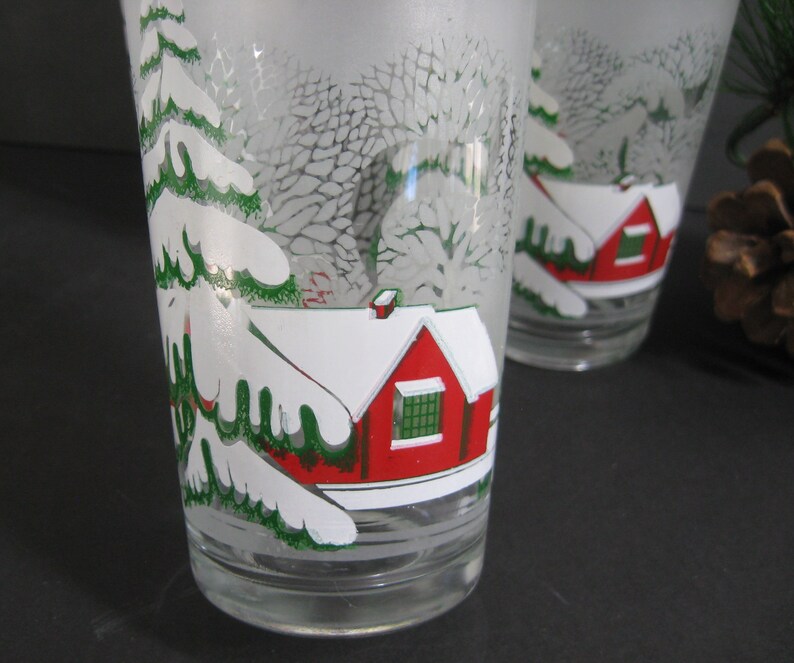 May include: Two frosted glass tumblers with a winter scene design. The scene features a red house with a white roof and a green tree covered in snow.