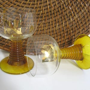 K&ouml;nnte beinhalten: Zwei Weingl&auml;ser aus bernsteinfarbenem Glas mit goldenen Akzenten. Die Gl&auml;ser haben einen breiten Kelch und einen hohen, gedrehten Stiel.