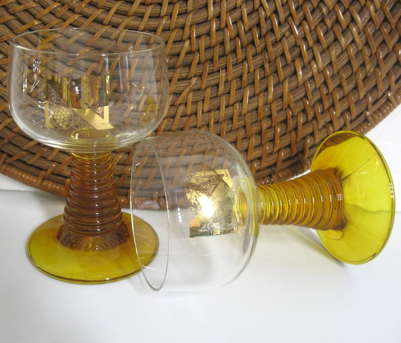 K&ouml;nnte beinhalten: Zwei Weingl&auml;ser aus bernsteinfarbenem Glas mit goldenen Akzenten. Die Gl&auml;ser haben einen breiten Kelch und einen hohen, gedrehten Stiel.