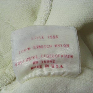 Op de afbeelding: Wit stoffen label met rode tekst. Het label luidt "Style 7556 100% Stretch Nylon Exclusive of Decoration RN 16942 Made in U.S.A."