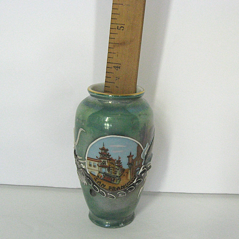 Pode incluir: Um pequeno vaso de cer&acirc;mica verde com uma cena pintada de Chinatown em S&atilde;o Francisco. O vaso tem uma borda decorativa prateada e uma r&eacute;gua de madeira no interior. O vaso tem aproximadamente 10 cm de altura.