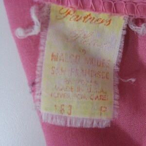 Puede incluir: Una etiqueta de ropa rectangular de tela blanca cosida sobre una tela rosa. La etiqueta presenta texto naranja que dice Partners by Malco Modes San Francisco, Made in U.S.A., e incluye instrucciones de cuidado, el n&uacute;mero 163 y la letra P.
