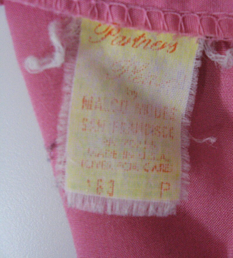 Puede incluir: Una etiqueta de ropa rectangular de tela blanca cosida sobre una tela rosa. La etiqueta presenta texto naranja que dice Partners by Malco Modes San Francisco, Made in U.S.A., e incluye instrucciones de cuidado, el n&uacute;mero 163 y la letra P.