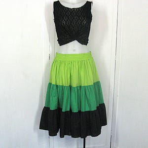 Op de afbeelding: Een tweedelige outfit met een zwarte kanten crop top en een gelaagde rok. De rok heeft drie horizontale kleurstroken: limoengroen, middengroen en zwart. De top is mouwloos en heeft een gedraaide voorkant.