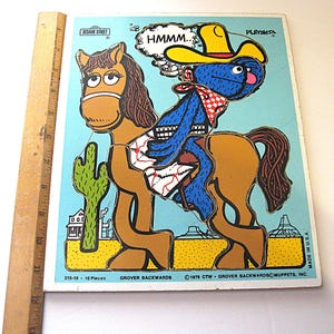 Pode incluir: Um quebra-cabe&ccedil;a Sesame Street Playskool com um personagem azul usando um chap&eacute;u de cowboy amarelo montado em um cavalo marrom em um deserto com um cacto. Uma r&eacute;gua de madeira &agrave; esquerda mostra uma altura de cerca de 33 cm.