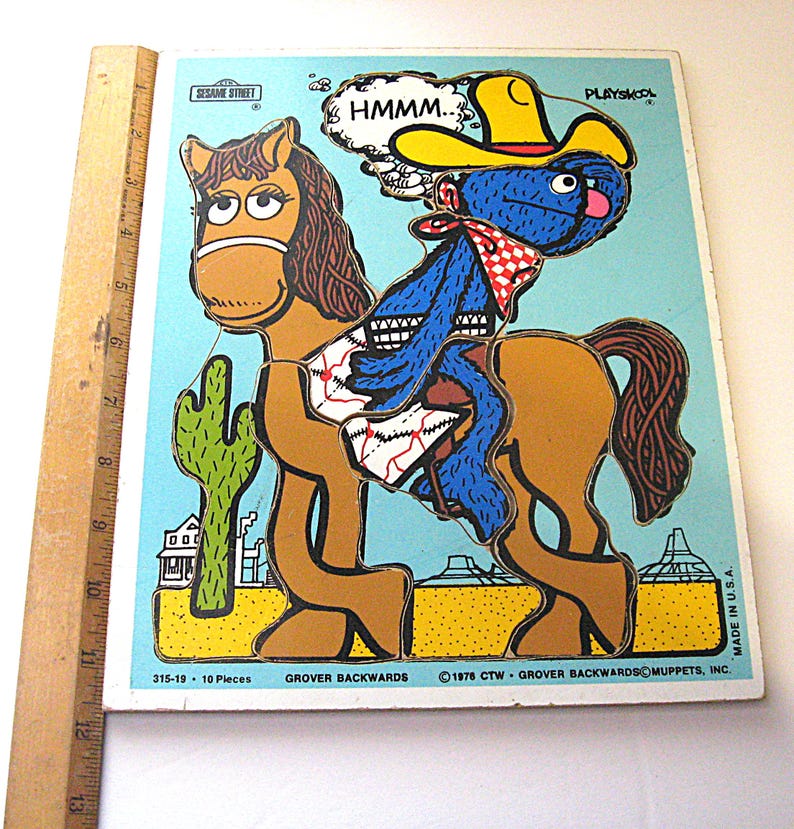 Pode incluir: Um quebra-cabe&ccedil;a Sesame Street Playskool com um personagem azul usando um chap&eacute;u de cowboy amarelo montado em um cavalo marrom em um deserto com um cacto. Uma r&eacute;gua de madeira &agrave; esquerda mostra uma altura de cerca de 33 cm.