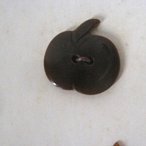 May include: A dark brown button shaped like an apple with a stem at the top. The button features a smooth, glossy surface and a single central hole for attachment, capturing the distinct rounded silhouette and leaf detail of the fruit design.