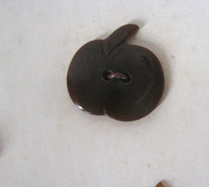 May include: A dark brown button shaped like an apple with a stem at the top. The button features a smooth, glossy surface and a single central hole for attachment, capturing the distinct rounded silhouette and leaf detail of the fruit design.