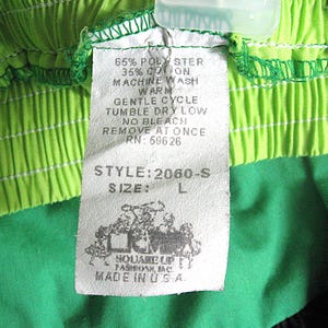 Op de afbeelding: Close-up van een kledinglabel met de tekst "65% POLY STER, 35% COTON, MACHINE WASH WARM, GENTLE CYCLE, TUMBLE DRY LOW, NO BLEACH, REMOVE AT ONCE, RN: 69626, STYLE: 2060-S, SIZE: L". Het label is bevestigd aan groene en limoengroene stof.