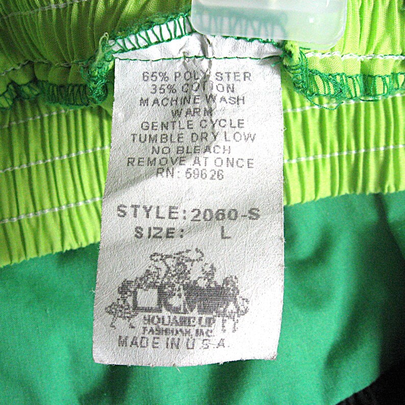 Op de afbeelding: Close-up van een kledinglabel met de tekst "65% POLY STER, 35% COTON, MACHINE WASH WARM, GENTLE CYCLE, TUMBLE DRY LOW, NO BLEACH, REMOVE AT ONCE, RN: 69626, STYLE: 2060-S, SIZE: L". Het label is bevestigd aan groene en limoengroene stof.