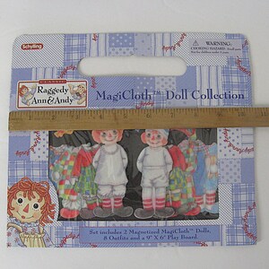 May include: A Raggedy Ann & Andy MagiCloth Doll Collection set. The package includes two magnetized dolls and eight outfits. The packaging is blue with a patchwork design and features the text "MagiCloth Doll Collection". A ruler measures 9 inches.