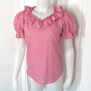 Puede incluir: Una blusa rosa de manga corta con un cuello ancho de volantes que enmarca el escote. La prenda presenta mangas abullonadas con pu&ntilde;os el&aacute;sticos de volantes y un dobladillo recto, confeccionada en un tejido liso y opaco de color s&oacute;lido.